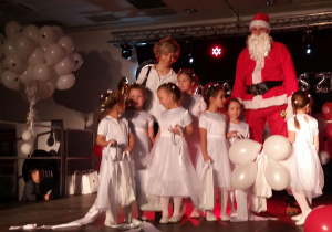 Na scenie stoją dziewczynki w białych sukienkach. Niektóre dziewczynki trzymają balony, inne szarfy. Na zdjęciu jest również nauczycielka i Mikołaj.